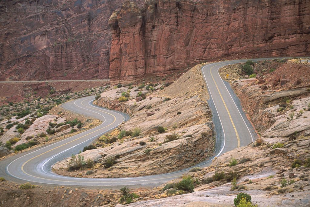 Photo picture of Two-lane paved road curves, twists, and turns on hill below red cliffsin Arches National Park, Utah