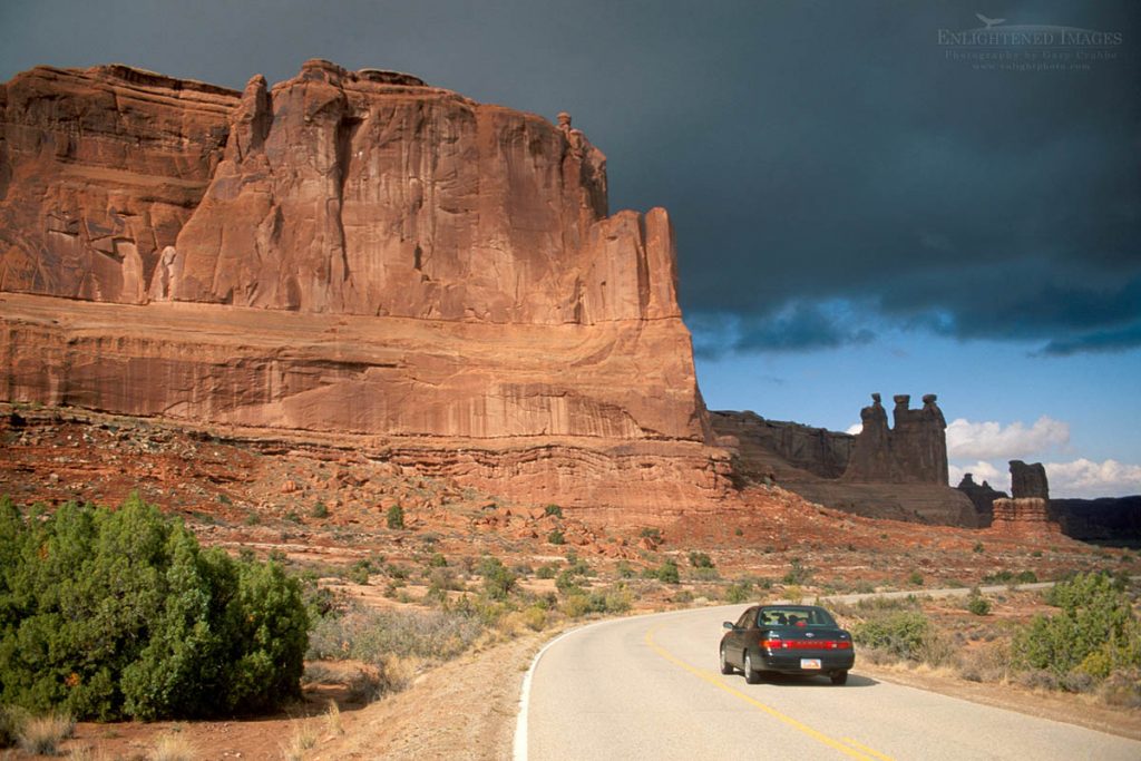 Photo picture of Car driving on two lane paved road below red rock cliffs, grey clouds, and blue sky, Arches National Park, Utah