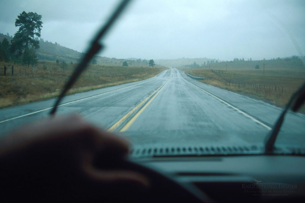 Photo picture of Hand on steering wheel and windshield wipers wiping while driving on empty two-lane road highway in a rainstorm, New Mexico